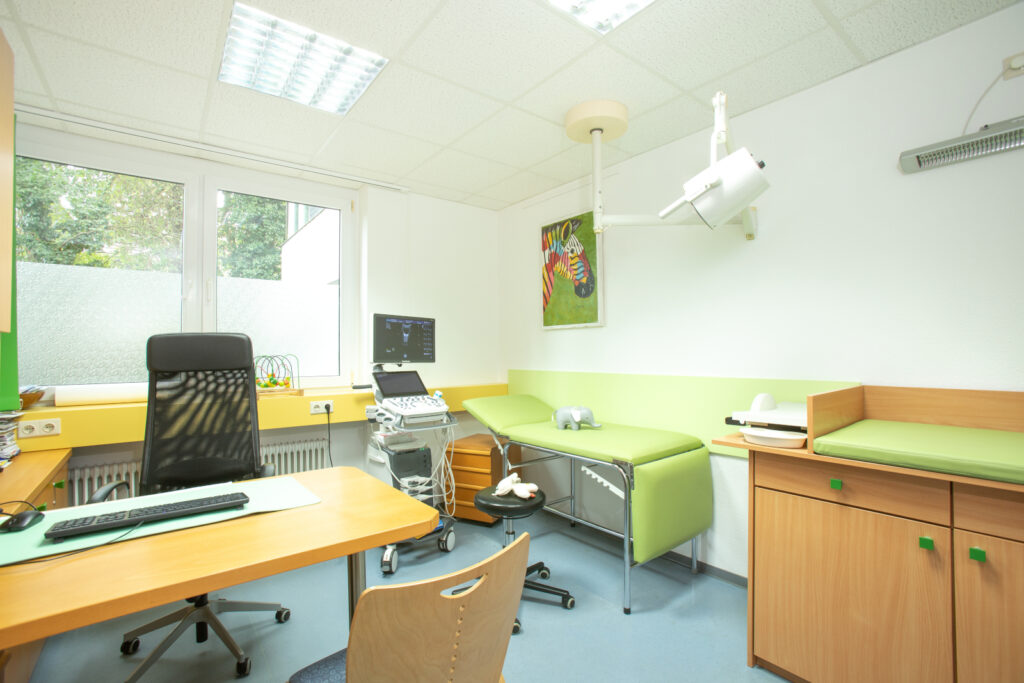 Einblick in ein Behandlungszimmer. Grün-Gelbes Dekor, sowie ein Schreibtisch und medizinische Geräte sind zu sehen.