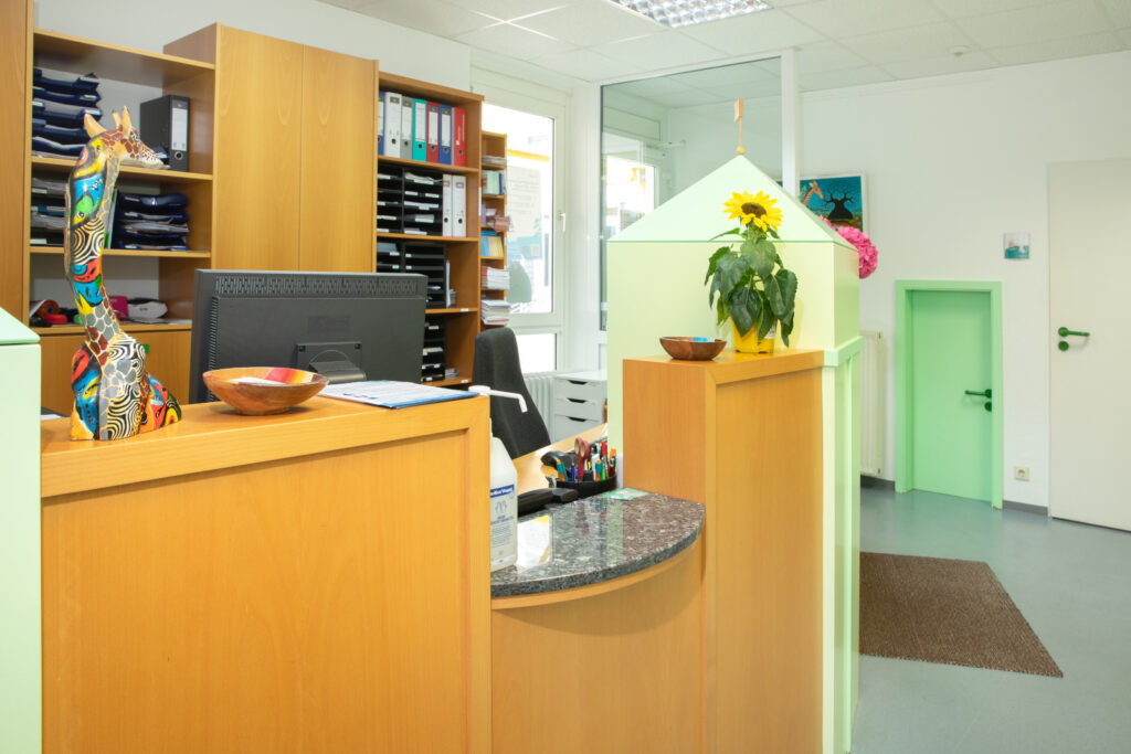 Eine Sicht auf den Empfangsbereich mit Schreibtisch, bunter Skulptur, Blumen und Gemälden.