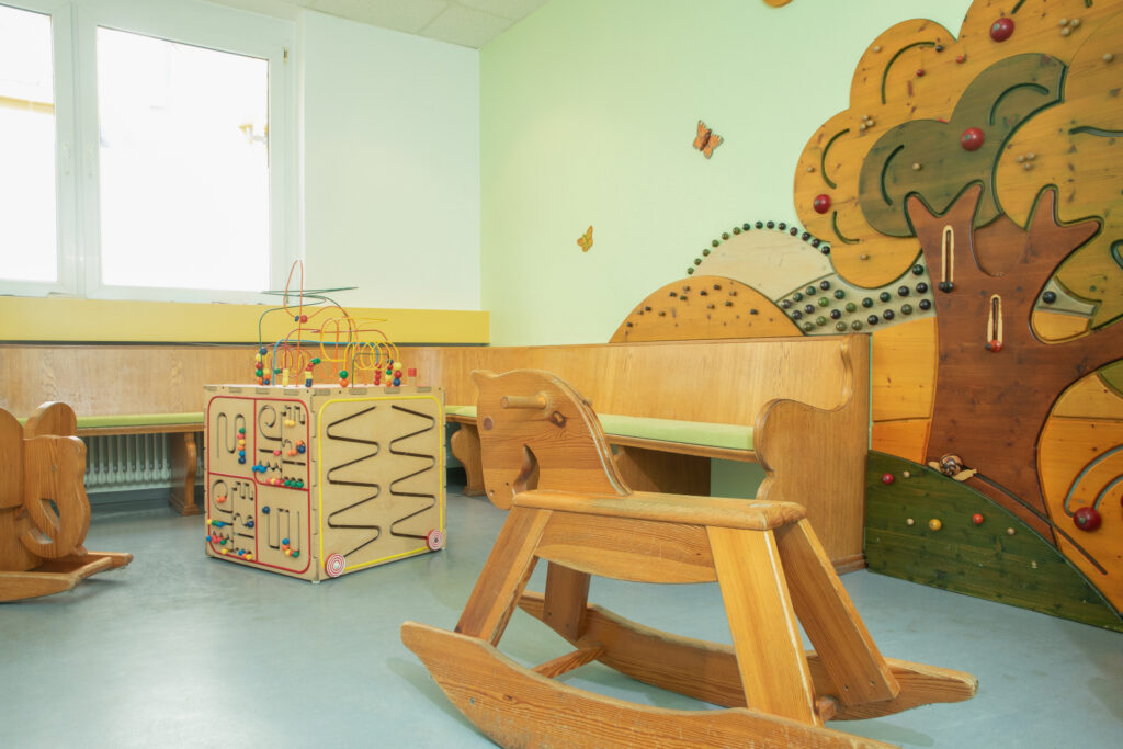 Bild eines Wartezimmers mit Holzspielzeugen, einer Spielwand und einem Schaukelpferd.